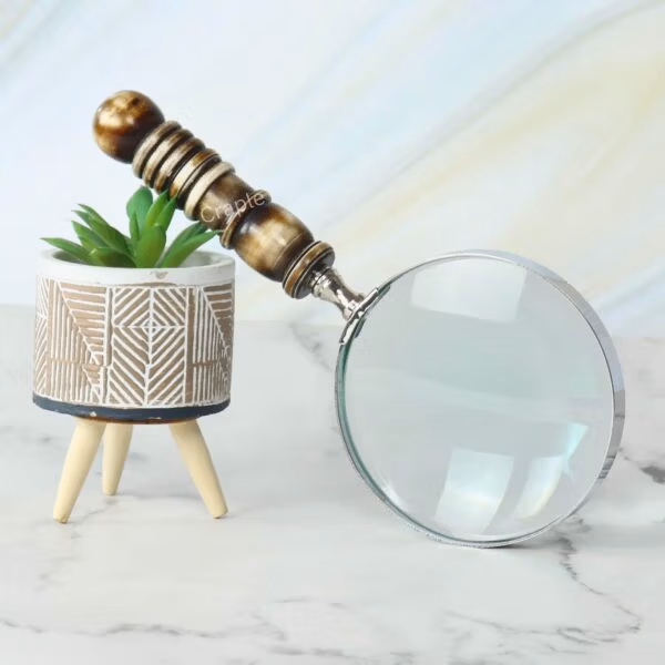 A decorative magnifying glass with a brown and white resin handle, propped against a stylish planter on a marble surface.