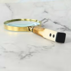 A decorative magnifying glass with a polished brass frame and a two-tone, domino-style handle lying on a marble surface.