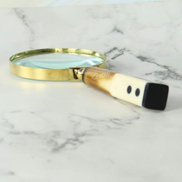 A decorative magnifying glass with a polished brass frame and a two-tone, domino-style handle lying on a marble surface.