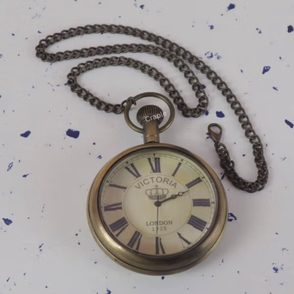 Victoria London 1915 antique brass pocket watch shown with its full fob chain laid out on a speckled surface.