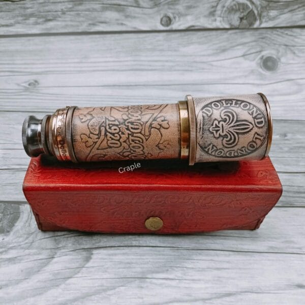 Vintage-style brass spyglass telescope with embossed leather, shown collapsed and resting on its red leather case.