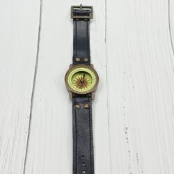 Full view of a vintage-style brass wrist compass with a long black leather strap, laid flat on a white wooden surface.