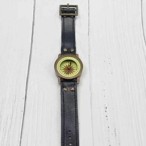 Full view of a vintage-style brass wrist compass with a long black leather strap, laid flat on a white wooden surface.