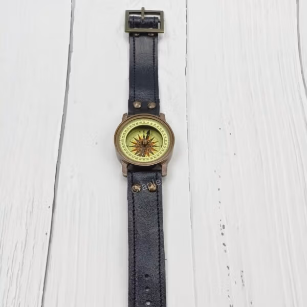 Full view of a vintage-style brass wrist compass with a long black leather strap, laid flat on a white wooden surface.