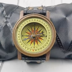 Antique brass wrist compass with a black leather strap, displayed on a black cushion for a product showcase.