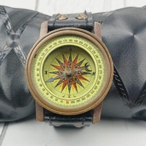 Antique brass wrist compass with a black leather strap, displayed on a black cushion for a product showcase.