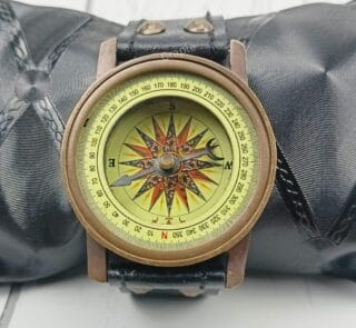 Antique brass wrist compass with a black leather strap, displayed on a black cushion for a product showcase.