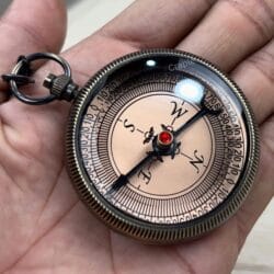 Angled view of the black antique brass compass held in hand, showing the glass cover and textured edge.