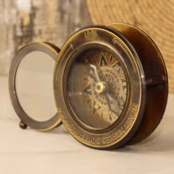 Vintage brass desk compass and paperweight with its sliding magnifier swung open, shown from an angled perspective.