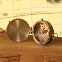 Angled view of the small brass pocket compass with its lid open, resting on a vintage map.