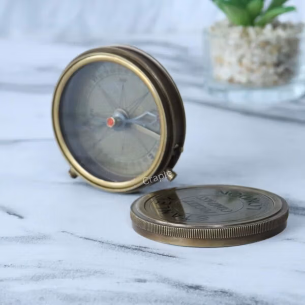 Antique brass pocket compass with its screw-off engraved lid placed beside it on a marble surface.