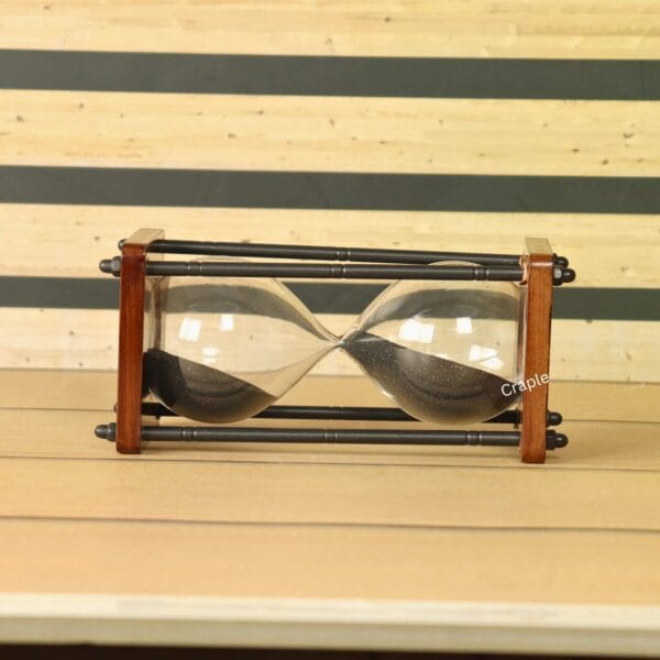 The wooden and antique brass hourglass resting horizontally on a wooden surface, showing the black sand inside.