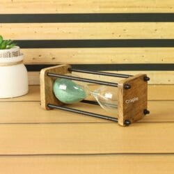 The wood and brass sand timer displayed as a decorative piece on a desk next to a small plant.