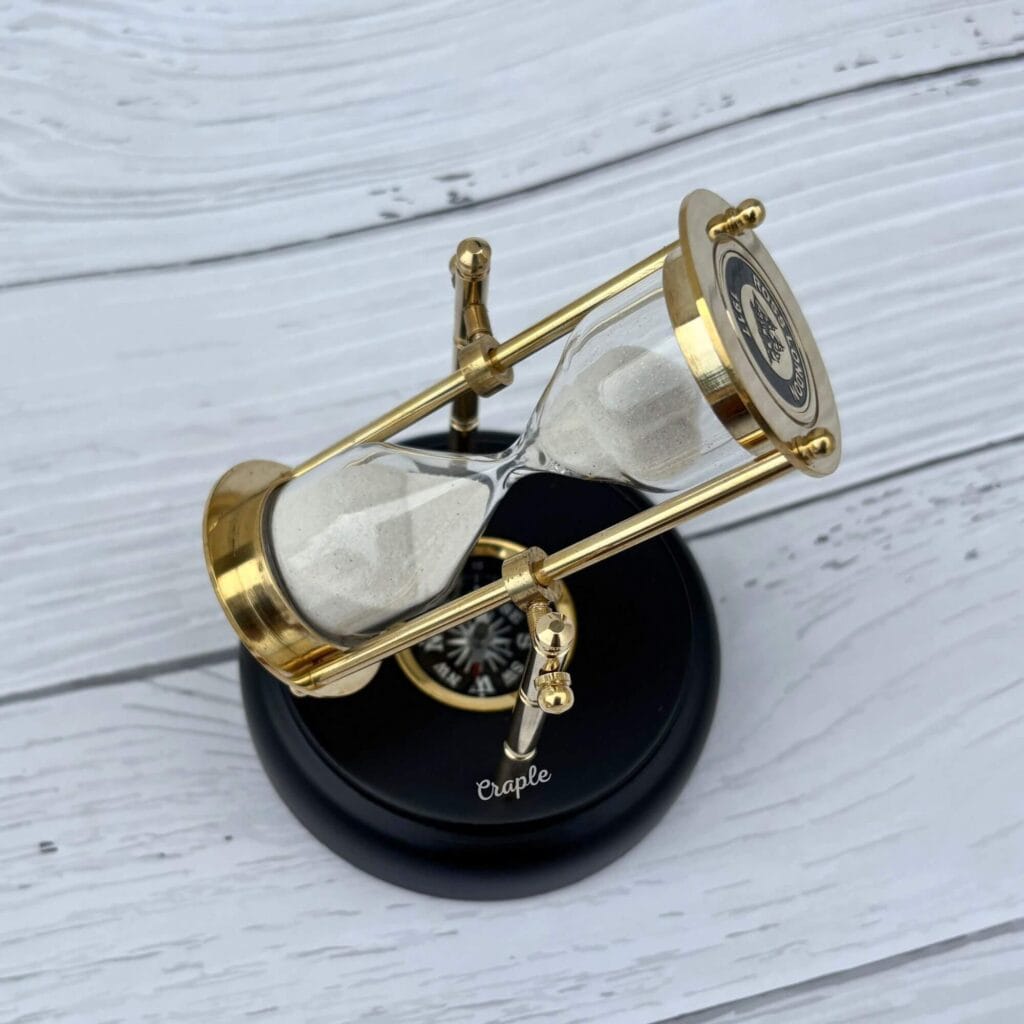 A rotating 3-minute hourglass with a polished brass frame and white sand, mounted on an engravable black wooden base with a built-in compass.