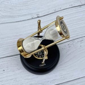 A rotating 3-minute hourglass with a polished brass frame and white sand, mounted on an engravable black wooden base with a built-in compass.