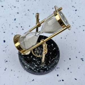 A rotating 3-minute brass hourglass with white sand, mounted on a black wooden base with a white speckled marble effect.