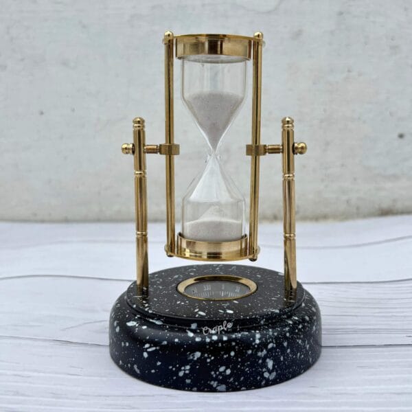 Front view of the brass hourglass in an upright position, showing the built-in compass embedded in the marble-effect wooden base.