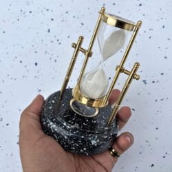 The brass and wood hourglass with a built-in compass held in a person's hand to show its size as a desktop accessory.