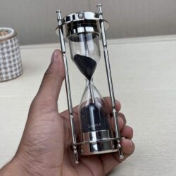 The nickel-finish hourglass and compass held in a hand to demonstrate its size and portability.