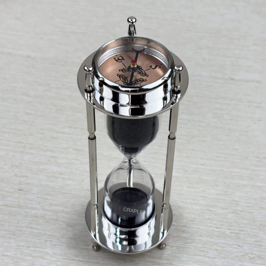 A multifunctional 3-in-1 desktop tool featuring a black sand hourglass, a compass, and a clock, all in a polished nickel-finish brass frame.