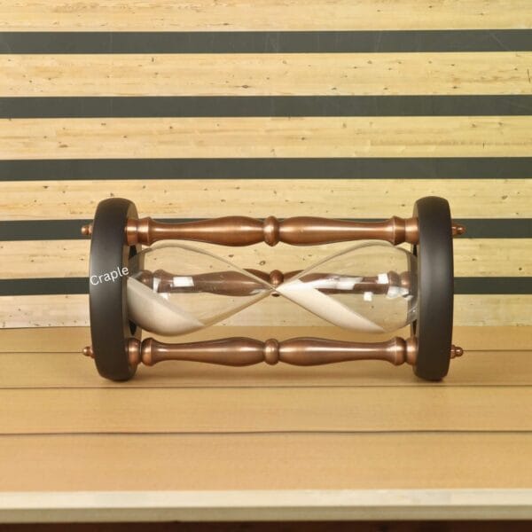 The antique copper and wood hourglass resting horizontally on a wooden surface, showing the classic pillar design.