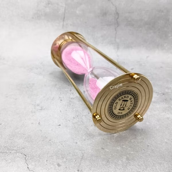 The 30-minute pink sand timer lying on its side, displaying the shiny brass pillars and engraved base.