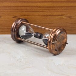 The decorative copper and black sand hourglass resting horizontally on a marble and wood surface.