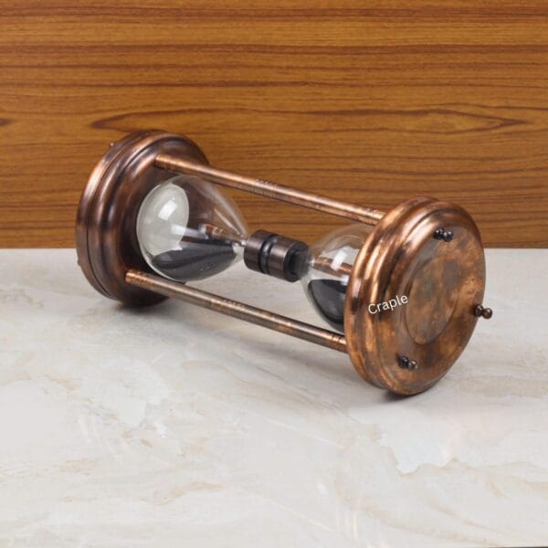 The decorative copper and black sand hourglass resting horizontally on a marble and wood surface.