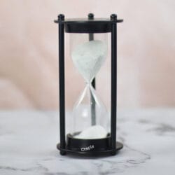 A modern 5-minute hourglass with a sleek black aluminum frame and fine white sand, sitting on a marble surface.