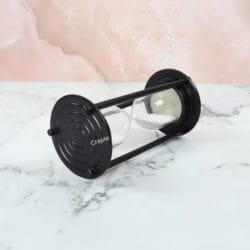 The black aluminum sand timer resting horizontally on a marble surface, showing the white sand settled in the glass.