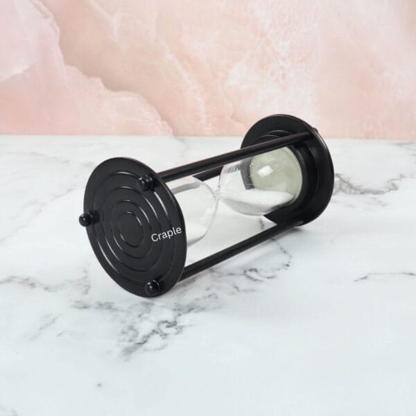 The black aluminum sand timer resting horizontally on a marble surface, showing the white sand settled in the glass.