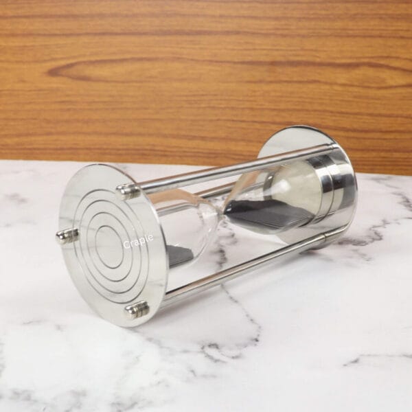 A modern 5-minute hourglass with a polished nickel finish and black sand, resting horizontally on a marble surface.