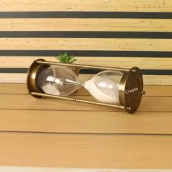 The antique brass one-hour sand timer resting horizontally on a wooden desk next to a small plant.