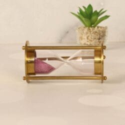 The 1-minute brass sand timer with pink sand resting horizontally next to a decorative plant.