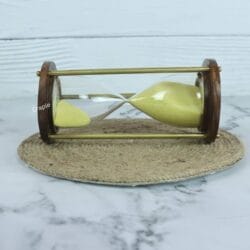 The wood and brass hourglass with yellow sand resting horizontally on a jute coaster.