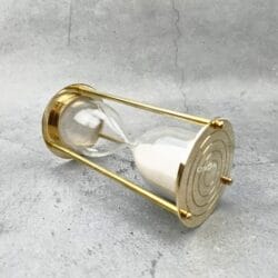 The polished brass sand timer resting horizontally on a stone surface, showing its pillar construction.