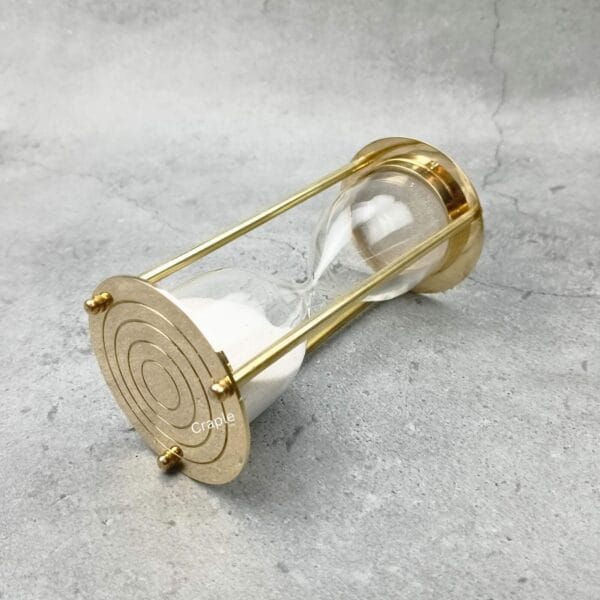 An angled side view of the brass hourglass lying down, highlighting the reflective finish of its base.