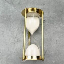 A close-up of the upper bulb of the brass hourglass, with white sand actively flowing through to the bottom.