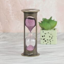 A vintage-style 1-minute hourglass with a cylindrical antique brass frame and sparkling pink sand.