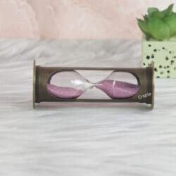 The cylindrical antique brass sand timer resting horizontally, showcasing the glass and pink sand inside.