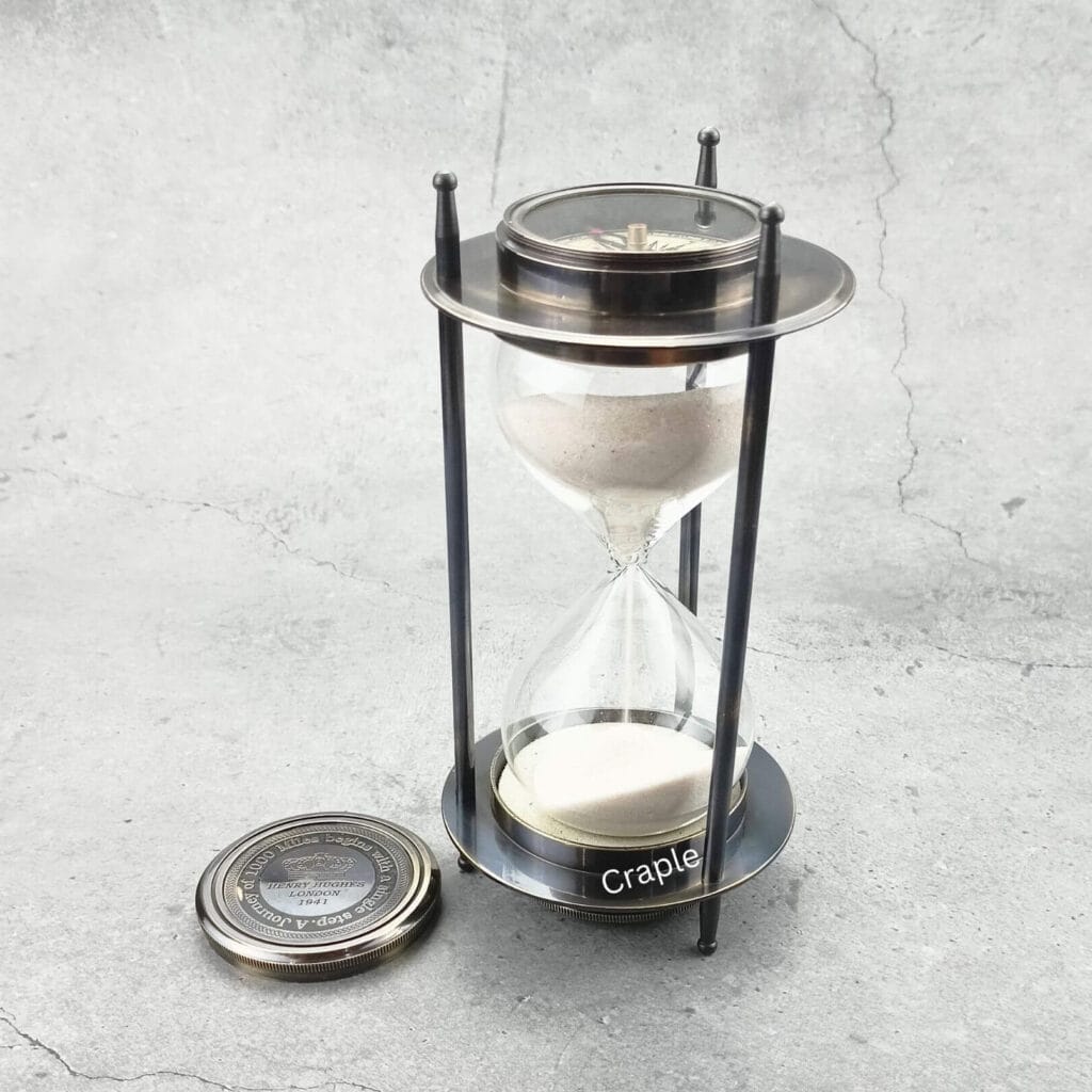 A vintage 5-minute hourglass with a black antique brass frame, white sand flowing, and a detachable compass.