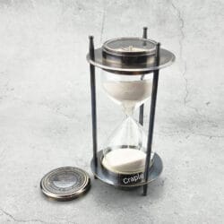 A vintage 5-minute hourglass with a black antique brass frame, white sand flowing, and a detachable compass.