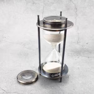 A vintage 5-minute hourglass with a black antique brass frame, white sand flowing, and a detachable compass.