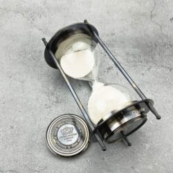 The dual compass sand timer tilted on its side, showing the glass hourglass and black antique finish.