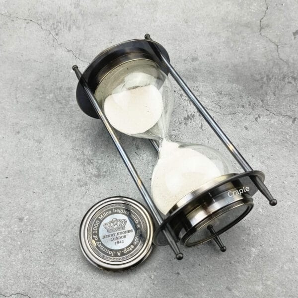 The dual compass sand timer tilted on its side, showing the glass hourglass and black antique finish.