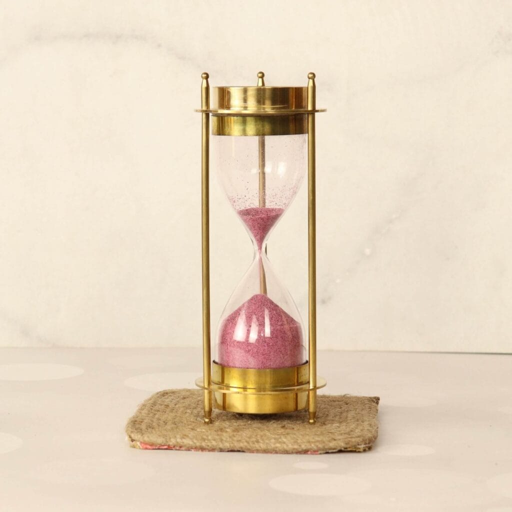 Shiny brass 5-minute hourglass sand timer with pink sand, standing vertically and showing the compass on top.