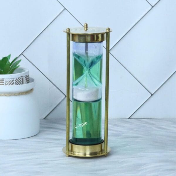 A modern liquid hourglass with a shiny brass frame and white sand suspended inside a green fluid-filled cylinder.