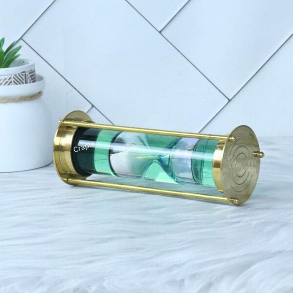 The brass and glass liquid sand timer resting horizontally, showing the inner hourglass floating in the green liquid.
