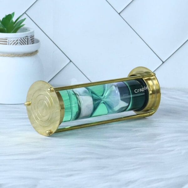 Angled view of the brass liquid timer on its side, highlighting the polished brass base with engraved circles.