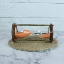 The wood and brass hourglass with orange sand resting horizontally on a jute coaster.